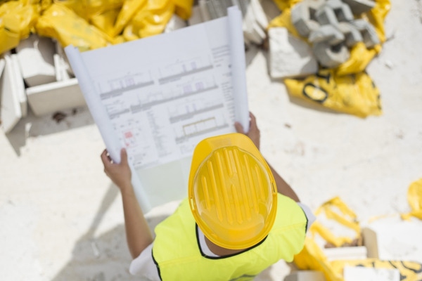 Builder in hardhat and safety vest looking at blueprint at construction site