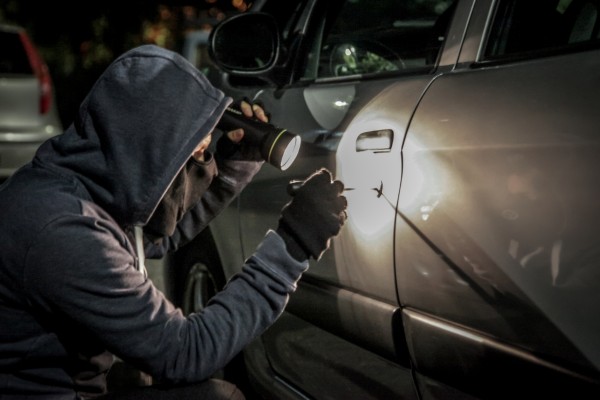 A man in hoodie and mask holding a flashlight and a lock picker trying to pick the lock of a car.