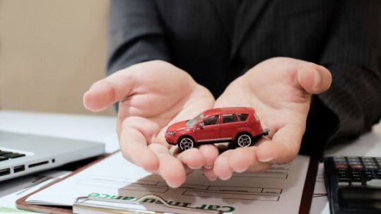 A red toy car being held in hand by a person, with car insurance form on the table.
